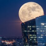 A large full moon rising over a modern glass office tower at night