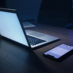 Half-closed laptop glowing in a dark office beside a phone, AI-generated illustration